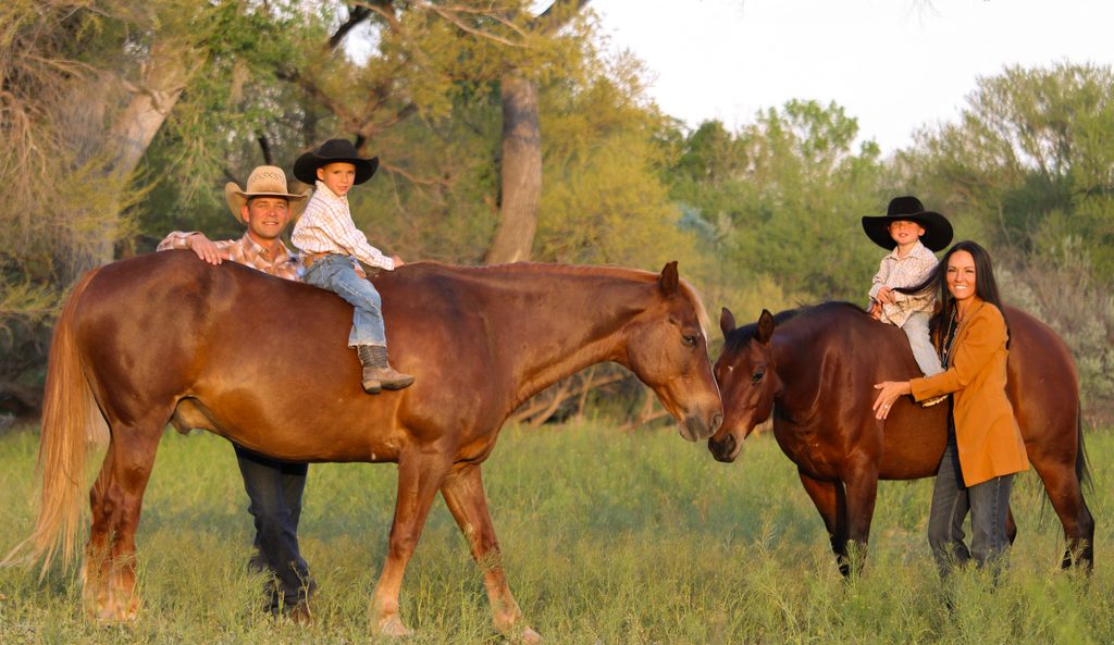 The Haught family with two young boys in black cowboy hats sitting on horses in a green pasture at sunset