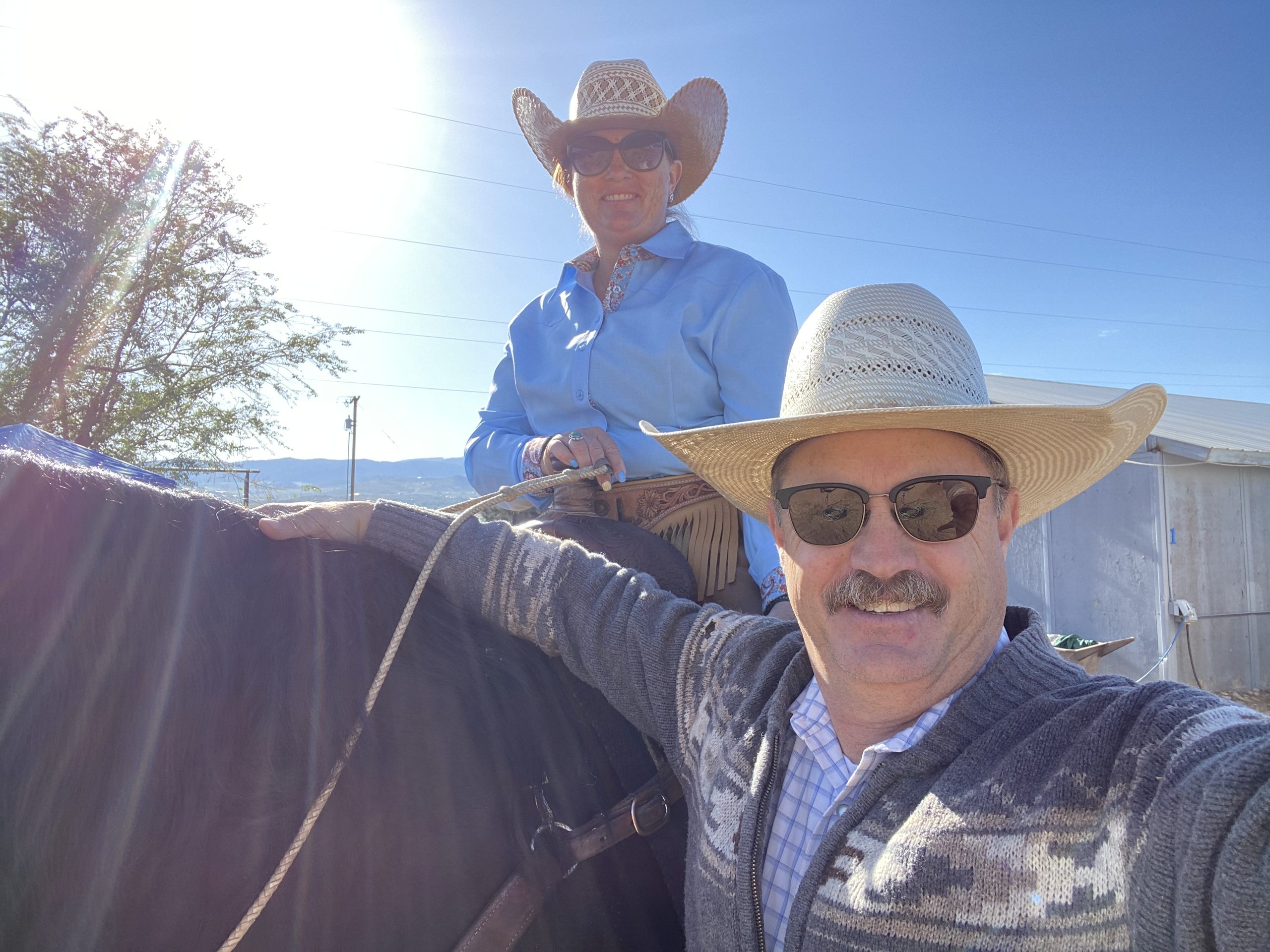 Smiling man in a straw cowboy hat taking a selfie beside his wife, who is mounted on a dark horse in a western show outfit
