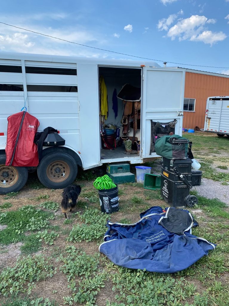 Open tack room of a white horse trailer with saddles, blankets, buckets, and gear spread across the grass while a dog walks by