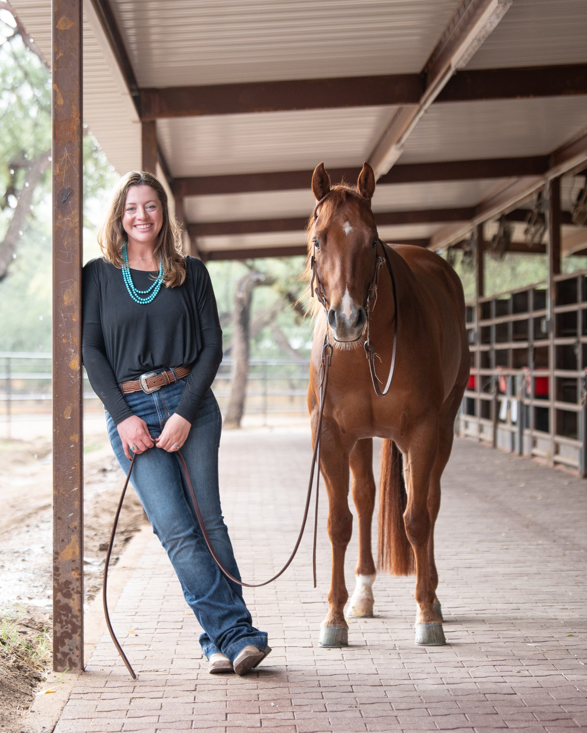 Caroline Arendt of Caroline Arendt Creative standing with her sorrel horse in a barn aisle.