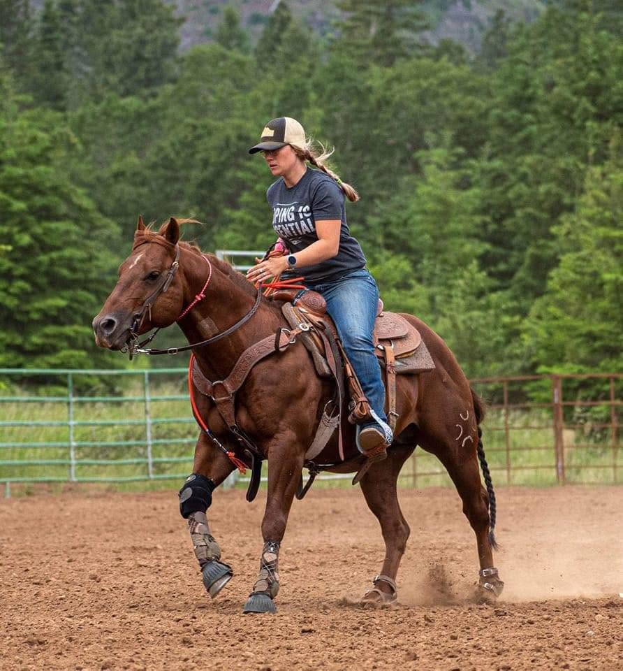 Jennifer Case riding a sorrel quarter horse in a dirt arena surrounded by pine trees