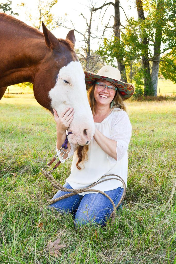 Jennifer Case laughing and hugging the face of a sorrel horse with a white blaze while kneeling in a grassy pasture